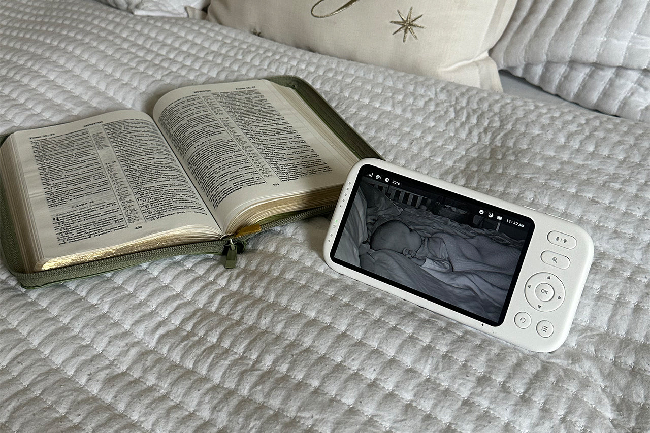 A baby monitor with a screen displaying a sleeping baby in night vision mode is placed next to an open Bible on a white quilted bedspread.