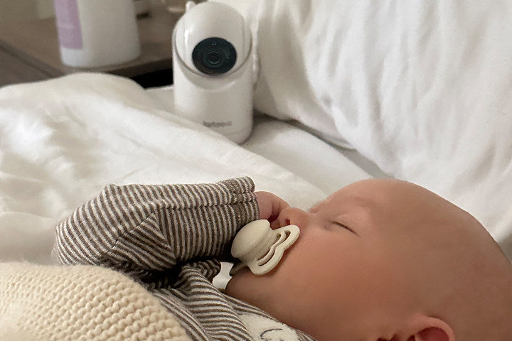 Close-up image of a sleeping baby on a white blanket, with a white baby monitor visible in the background. The baby is wearing a striped outfit and has a pacifier in its mouth, lying on its side with eyes closed, appearing peaceful.
